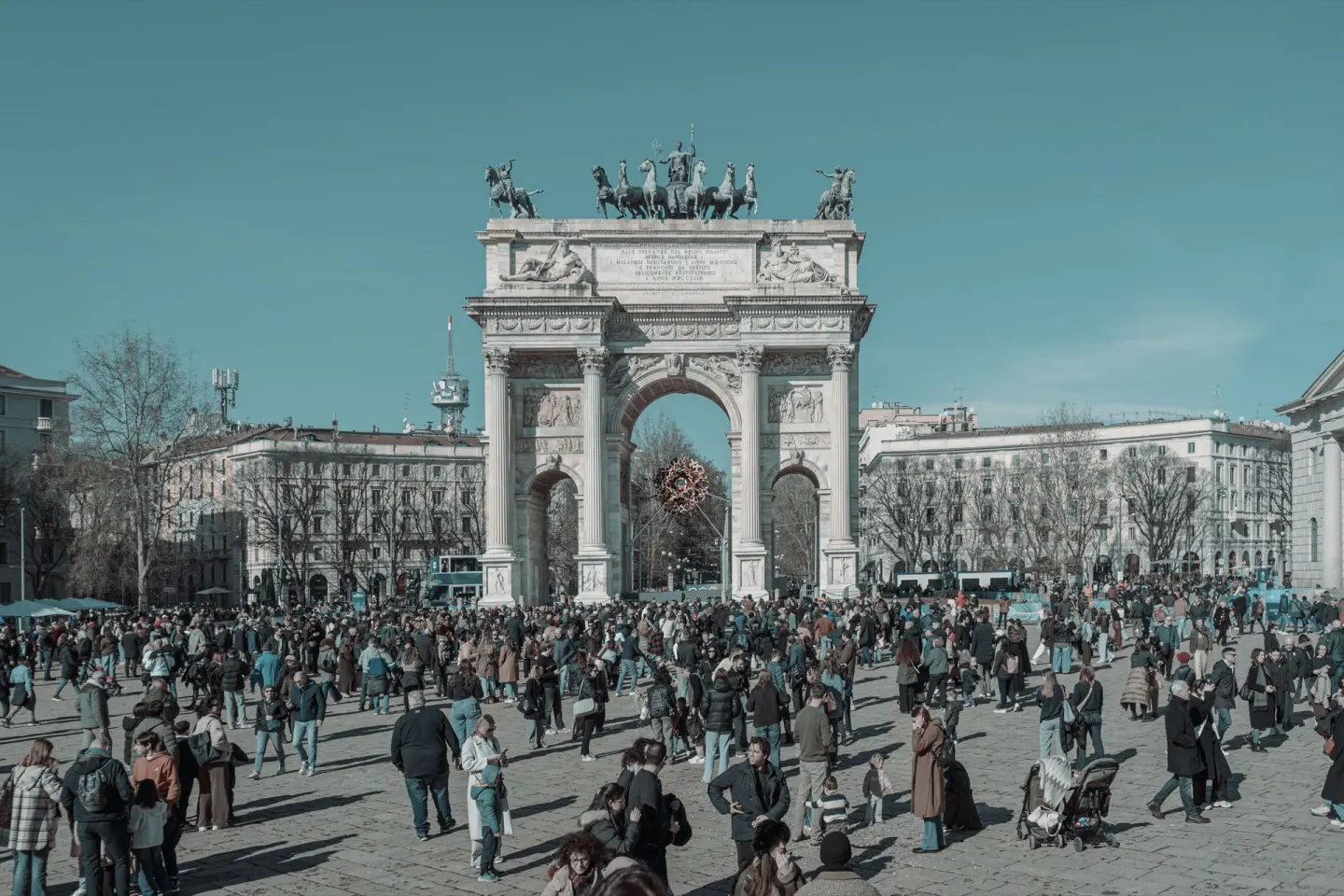 Gran arco triunfal neoclásico de mármol con columnas y una escultura de caballos y carro en lo alto, en una plaza llena de gente bajo un cielo azul, con edificios de fachadas claras a ambos lados