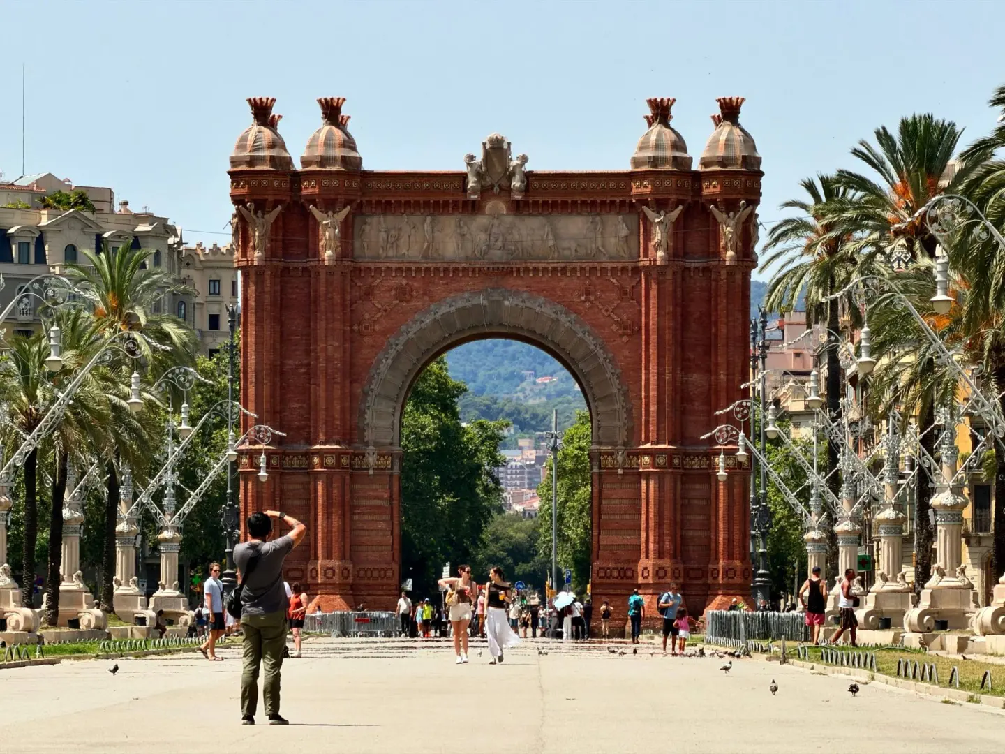 Arco de ladrillo rojo con relieves y torretas en un paseo con palmeras y farolas, con gente caminando