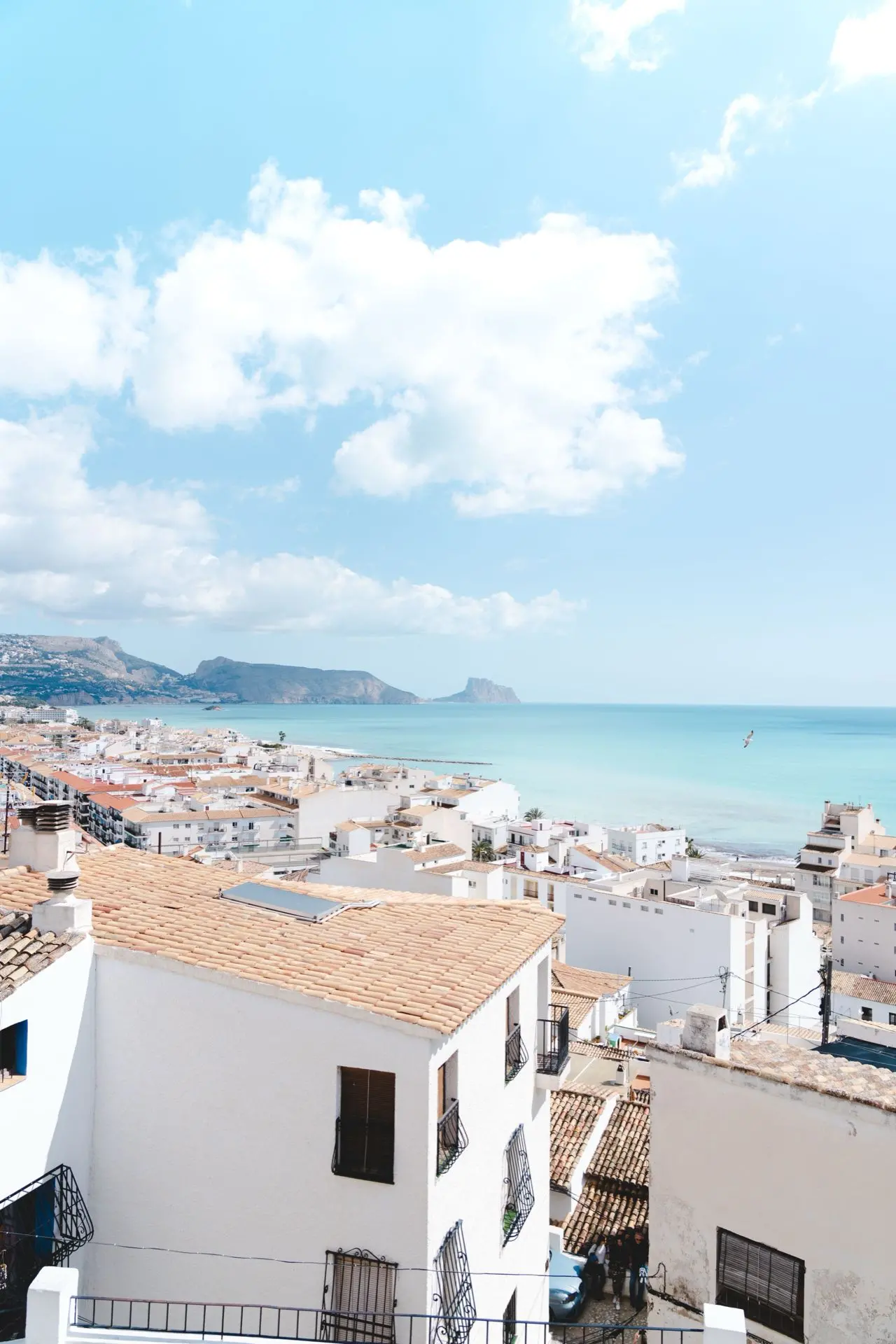 Vista desde lo alto de tejados de teja y casas blancas, con el mar azul turquesa y un gran peñón al fondo