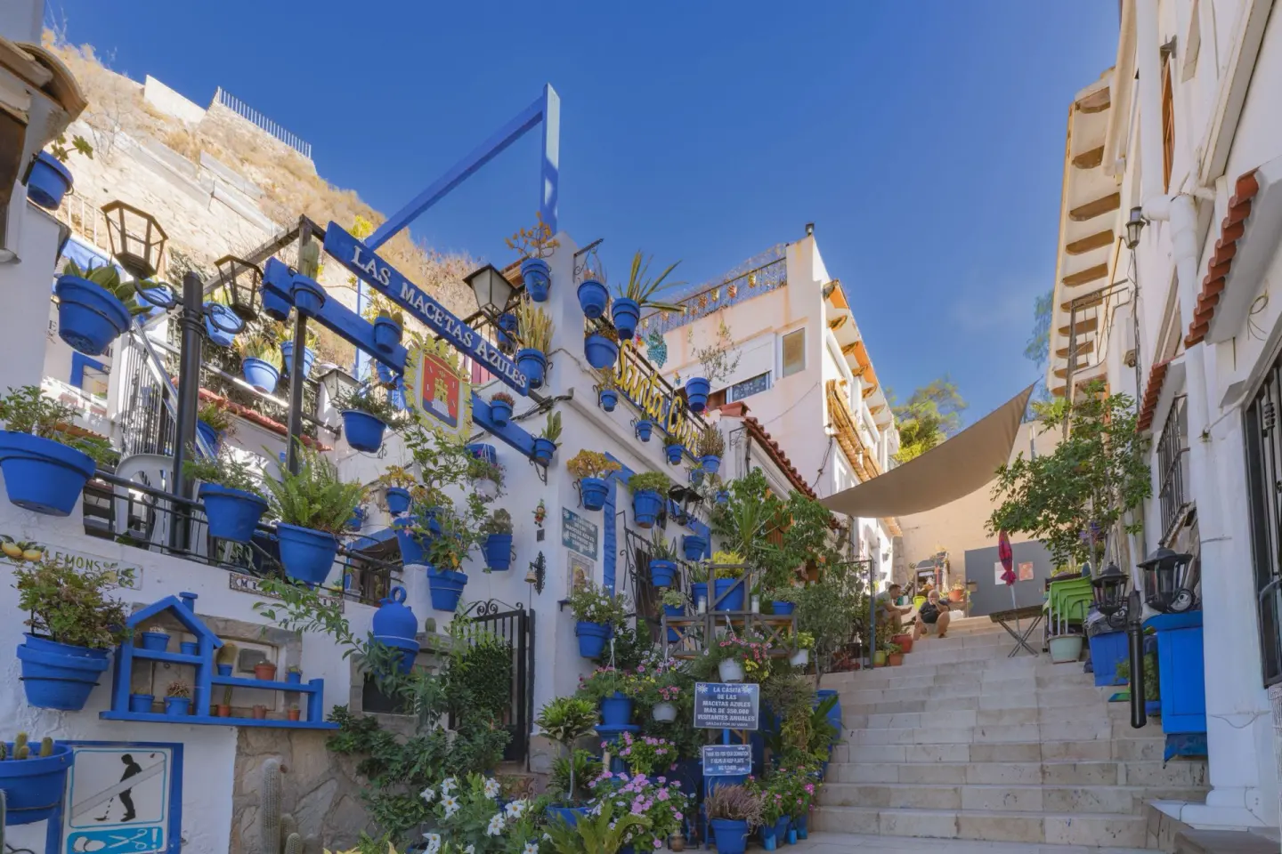 Calle empinada de casas blancas con escaleras y decenas de macetas azules colgadas en las fachadas, bajo un cielo azul