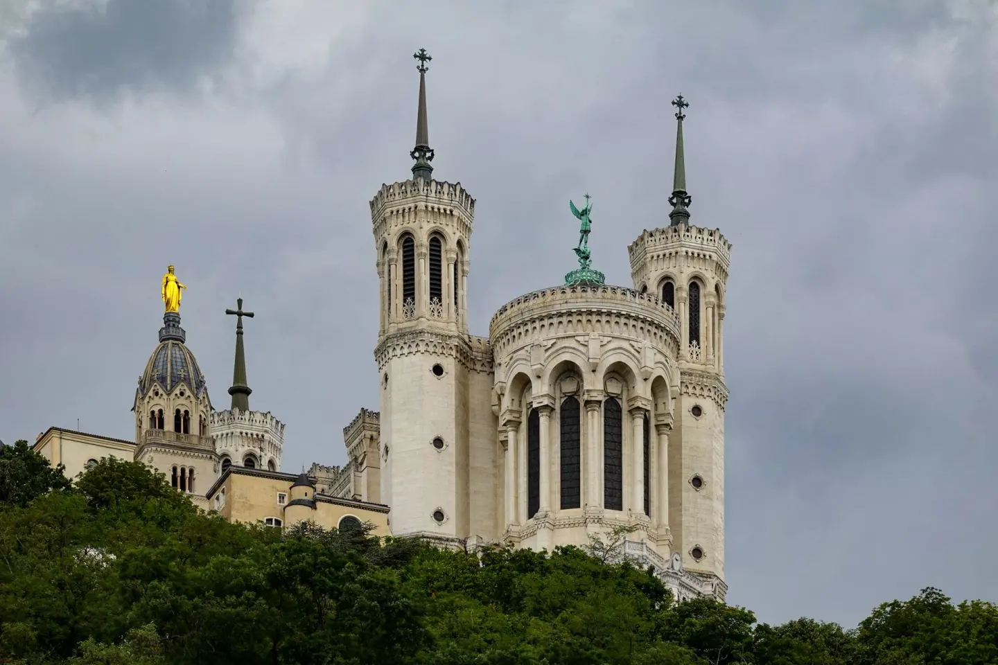 Torres y cúpulas de una gran basílica de piedra clara con una estatua dorada y otra verde en lo alto, sobre una colina de árboles, bajo un cielo de nubes oscuras
