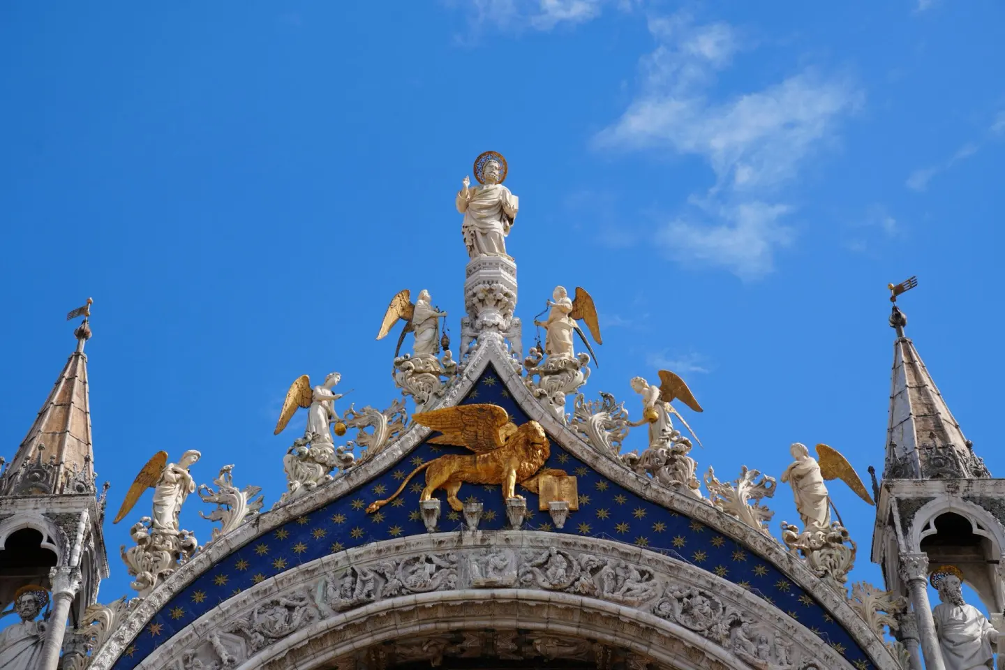 Remate de una fachada con un gran león alado dorado sobre fondo azul con estrellas doradas, rodeado de ángeles y estatuas de mármol blanco, bajo un cielo azul con nubes