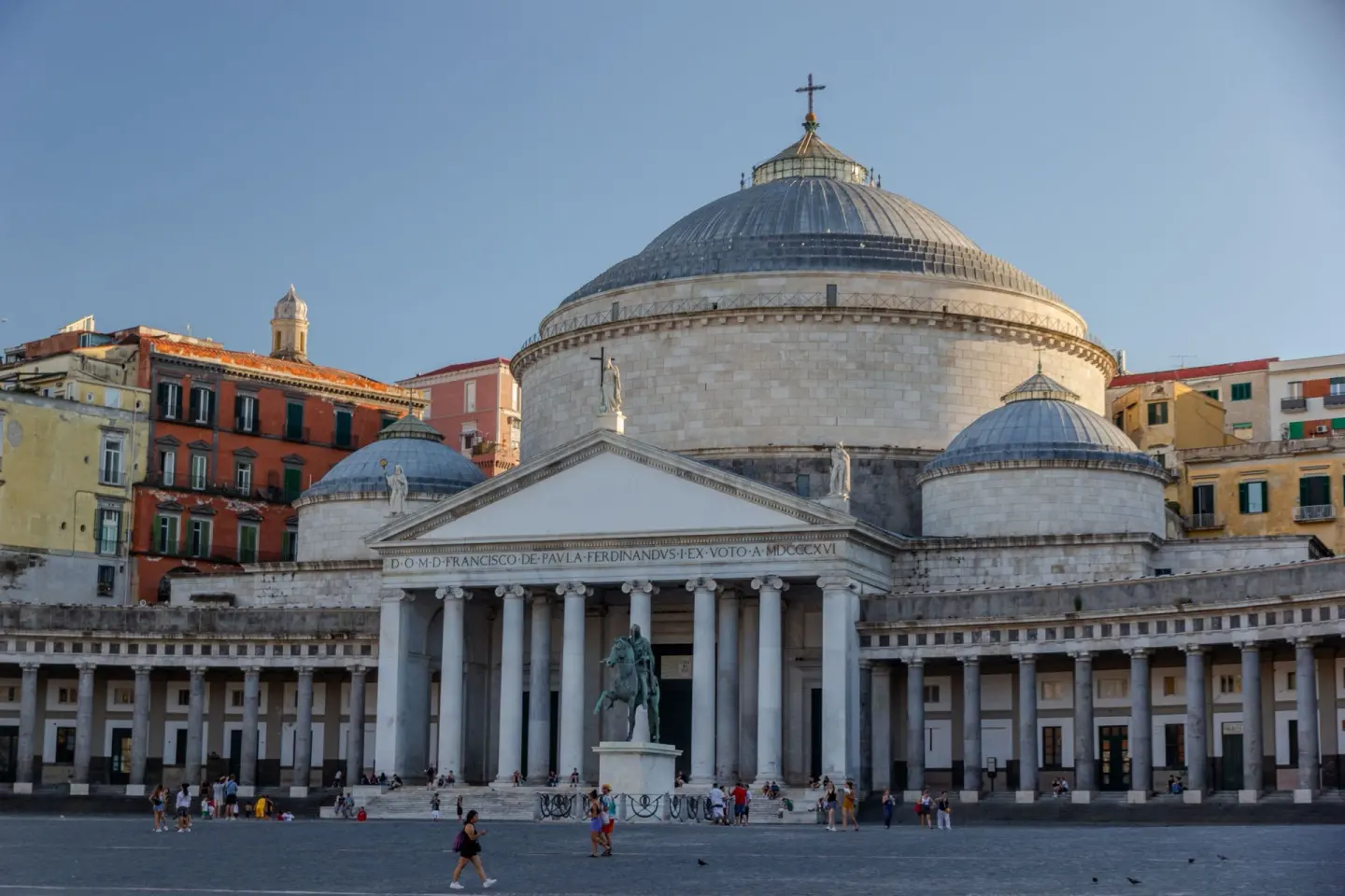 Iglesia neoclásica de gran cúpula gris con un pórtico de columnas y un pórtico semicircular de columnas a ambos lados, con una estatua ecuestre de bronce delante, en una gran plaza con gente y edificios de colores al fondo