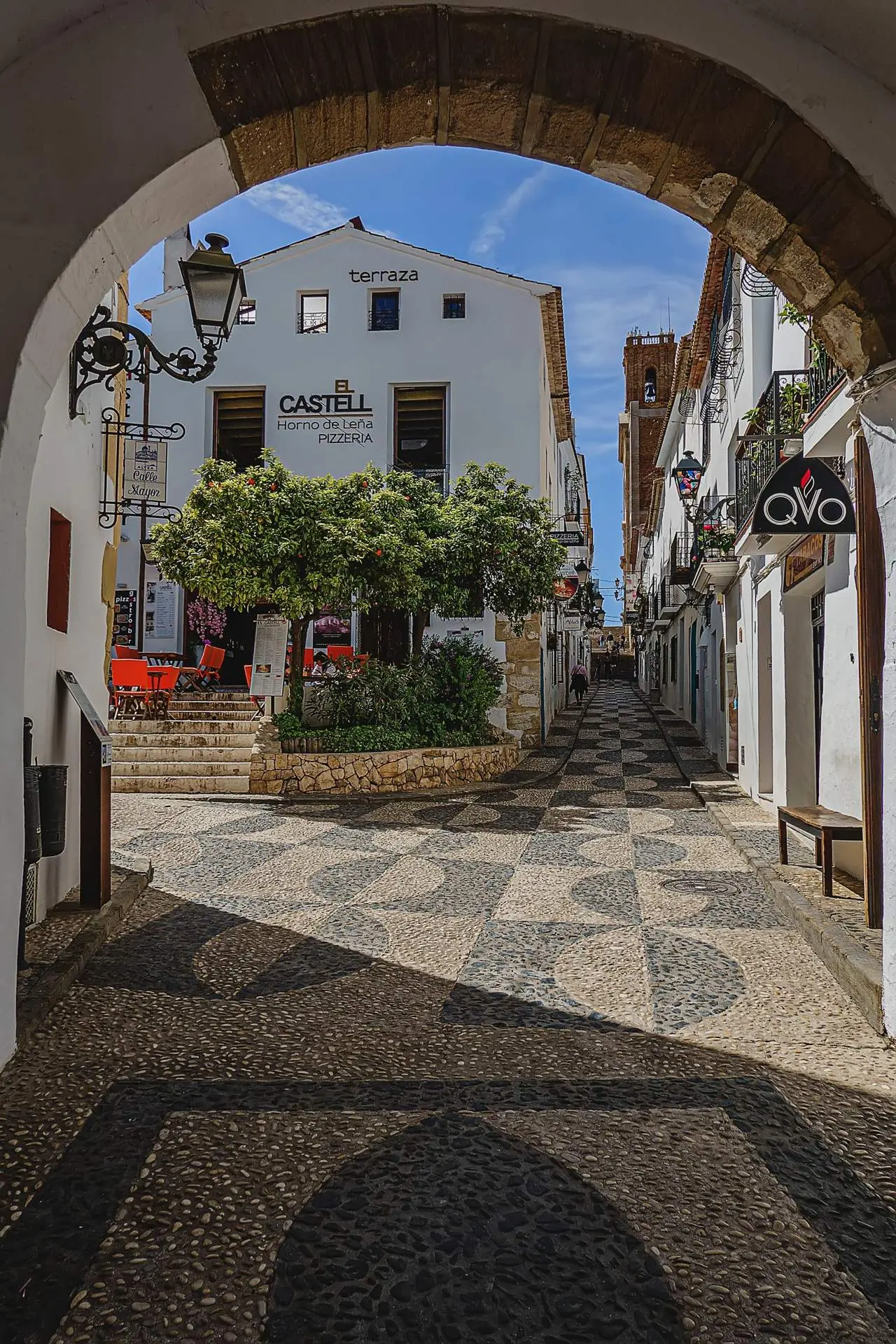 Calle con suelo de guijarros en mosaico vista a través de un arco, con un naranjo en una plaza y un campanario de ladrillo al fondo