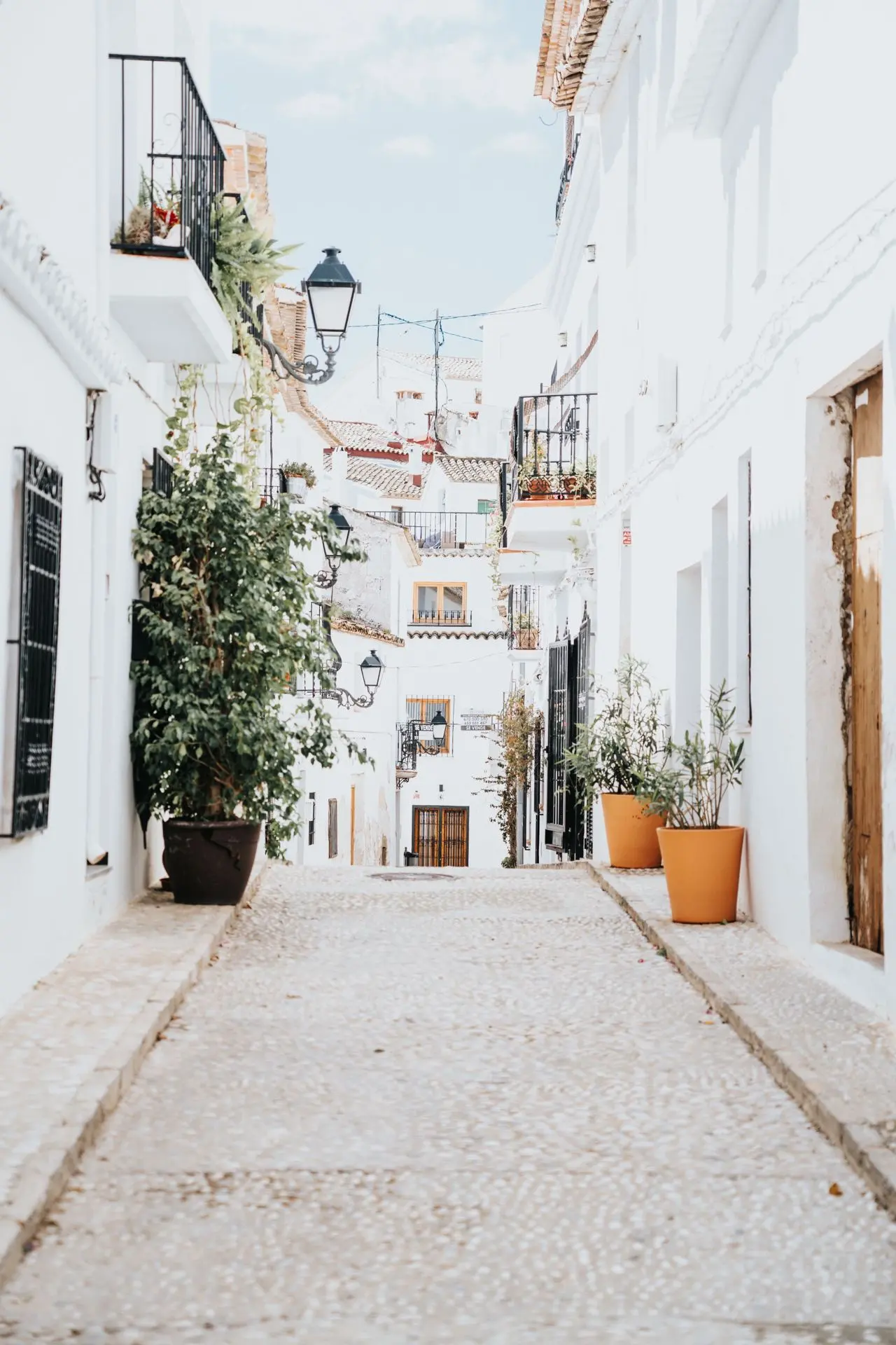 Calle empinada de casas blancas con macetas, farolas de forja y balcones, bajo un cielo claro