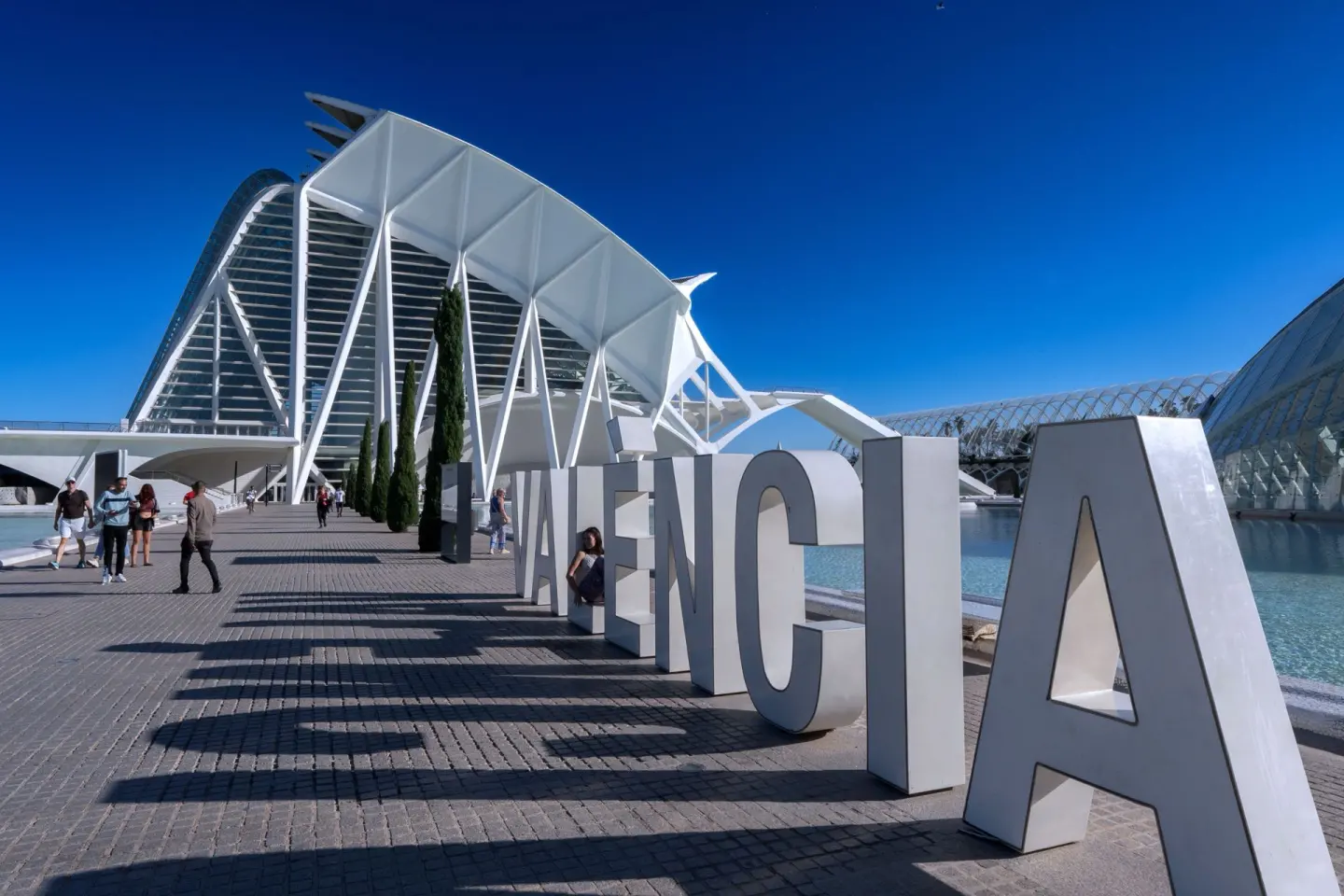 Letras gigantes blancas que forman la palabra «Valencia» junto a un estanque, con un edificio de estructura blanca al fondo y gente paseando