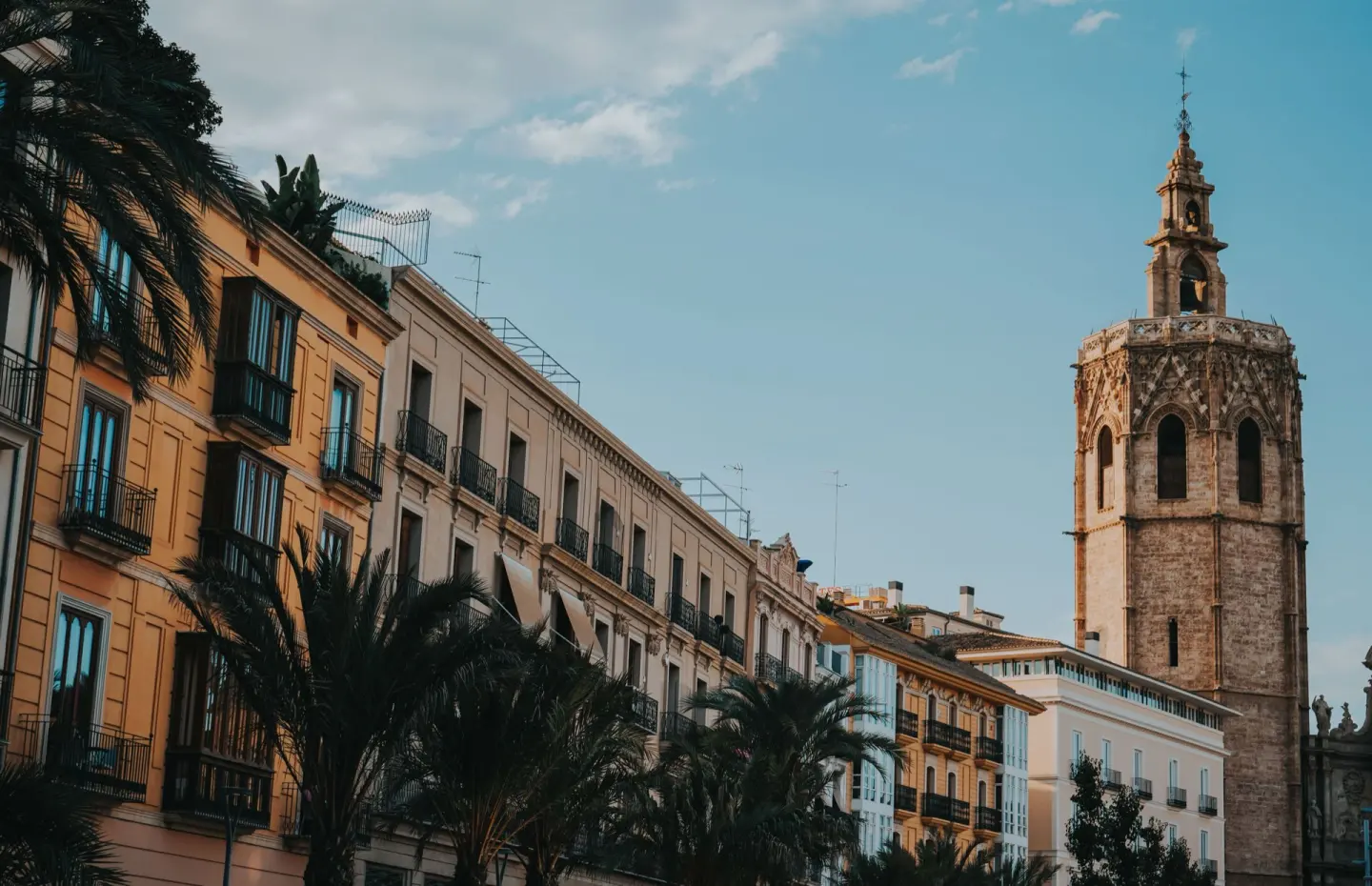 Torre campanario octogonal de piedra junto a edificios de fachadas amarillas y beis, con palmeras en primer plano