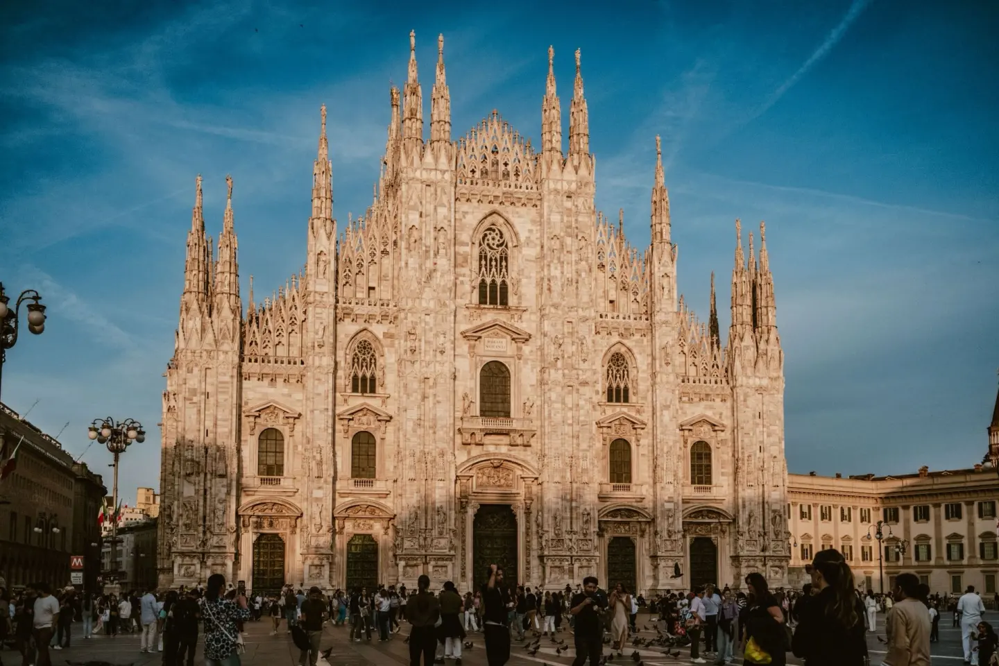 Fachada de una catedral gótica de mármol con numerosas agujas y estatuas, iluminada por la luz cálida del atardecer sobre un cielo azul, con gente y palomas en la plaza