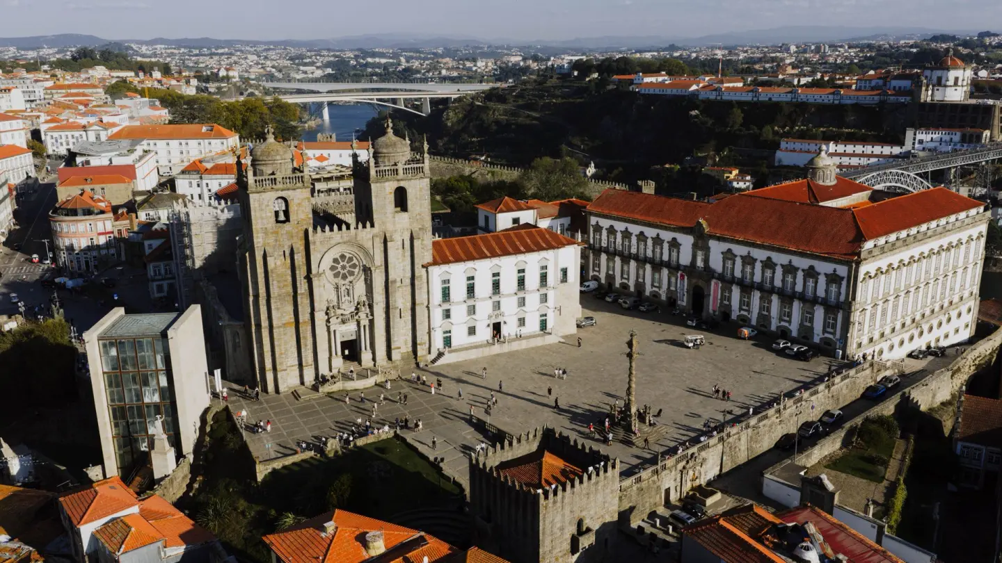 Vista aérea de una catedral de piedra con dos torres, rosetón y almenas, junto a una gran plaza empedrada con una columna en el centro y un palacio de fachada blanca con tejado rojo, con un río y un puente al fondo