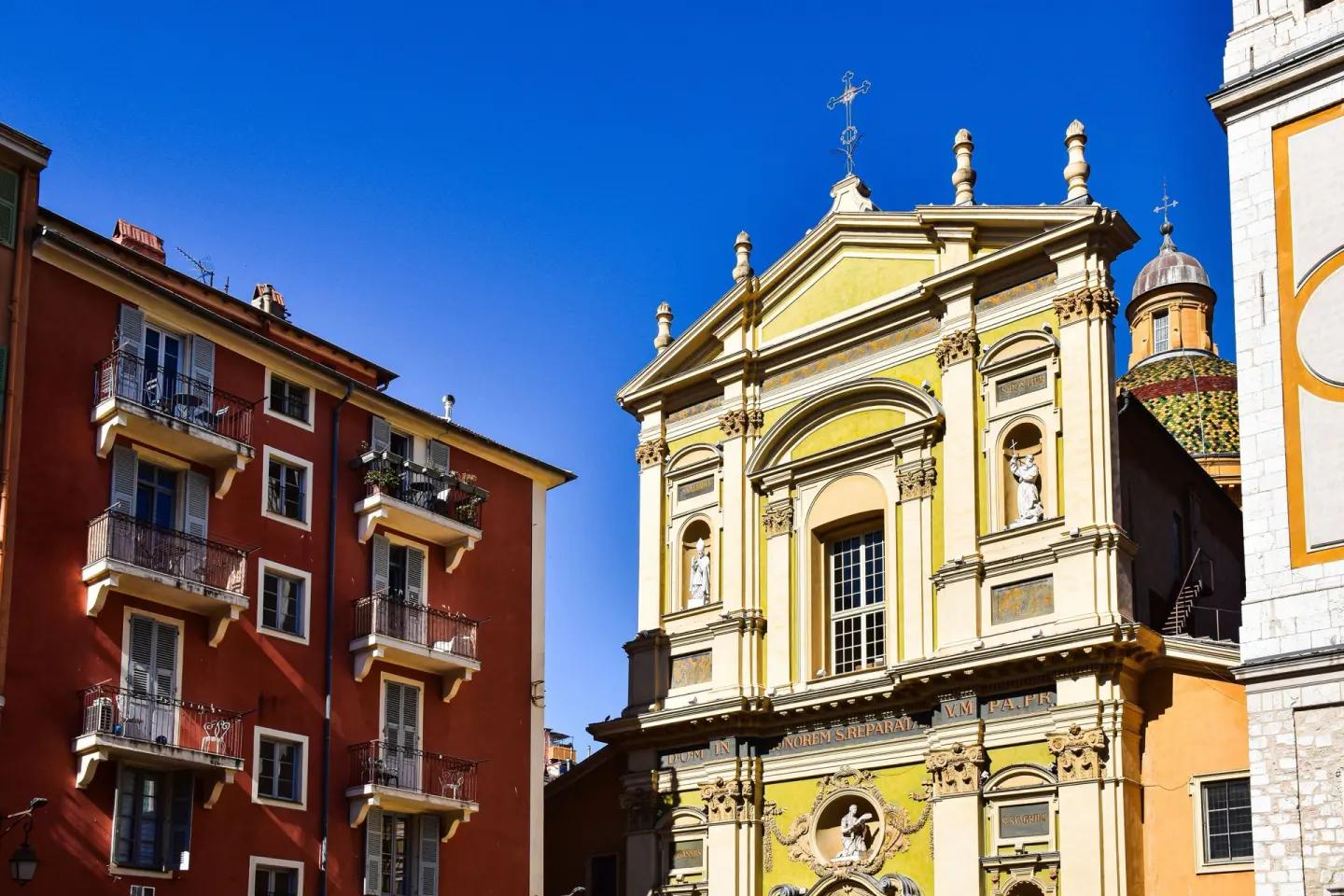Fachada barroca amarilla y crema de una iglesia con columnas y estatuas, junto a un edificio rojo de balcones y una cúpula de tejas de colores, bajo un cielo azul intenso