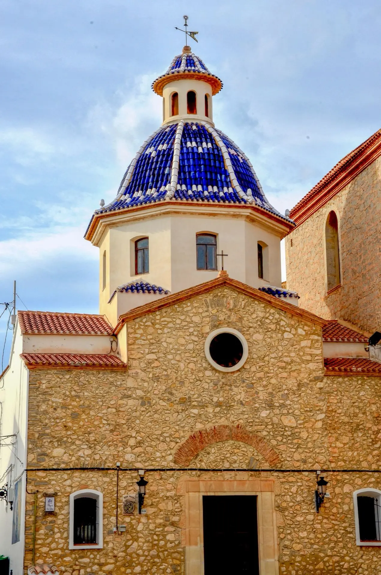 Iglesia de piedra con una cúpula cubierta de tejas azules y blancas sobre un cielo con nubes