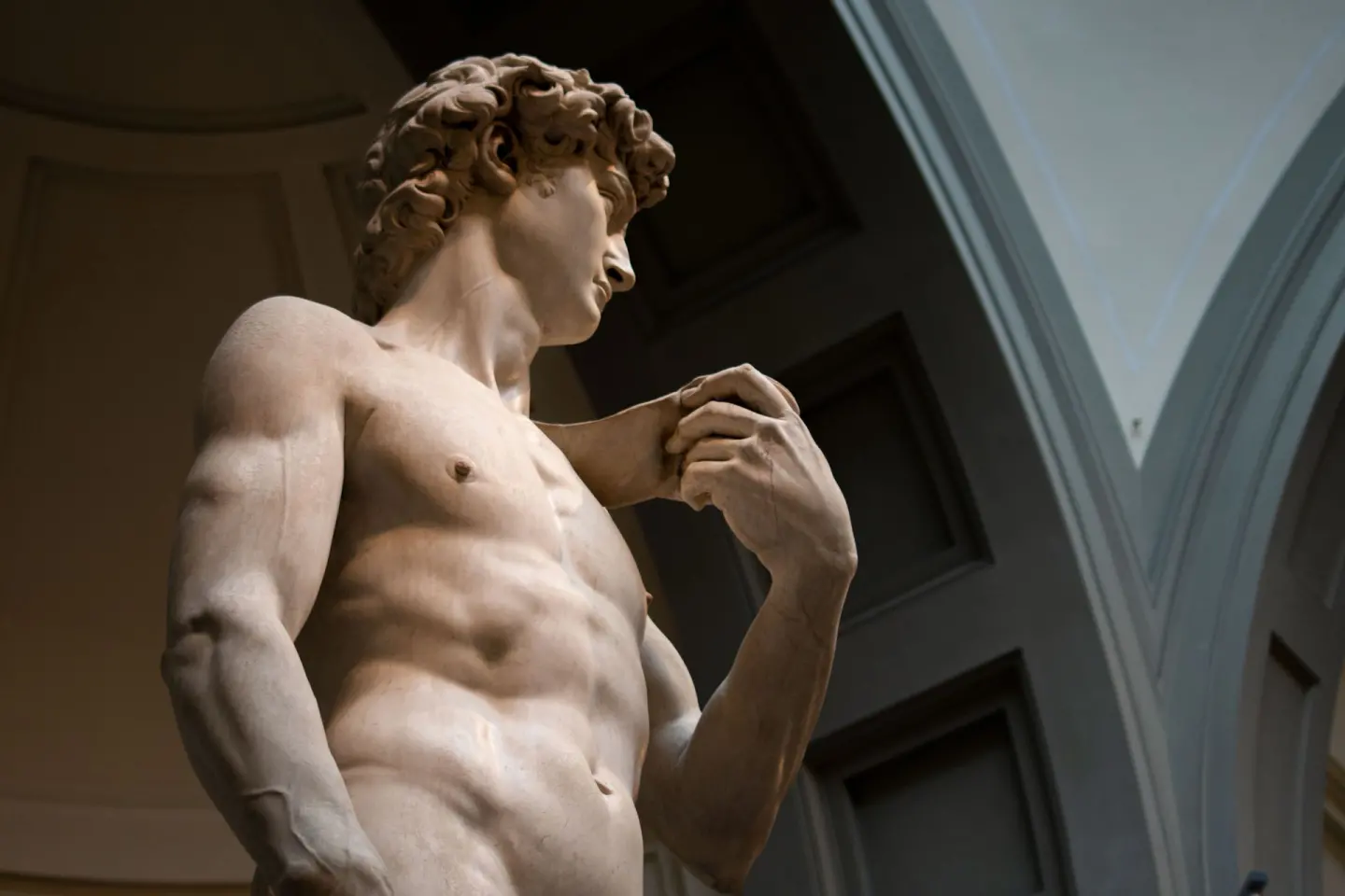 Escultura de mármol de un hombre joven desnudo de pelo rizado, vista desde abajo, con la mano izquierda levantada junto al hombro, en una sala de paredes oscuras con arcos