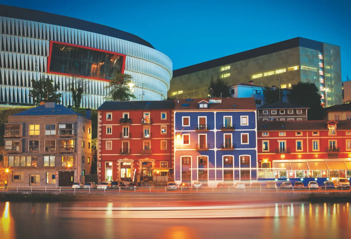 Estadio de fachada curva y metálica iluminada al anochecer, sobre casas de colores rojo y azul junto al agua, con luces que dejan estelas en la ría