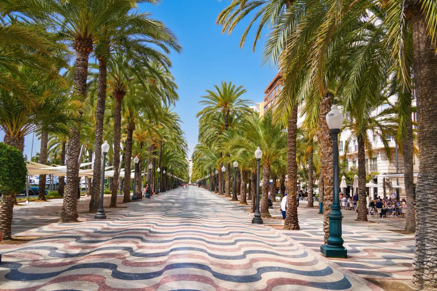 Paseo peatonal entre filas de palmeras y farolas, con un pavimento de mosaico ondulado en rojo, negro y blanco, y terrazas a la derecha