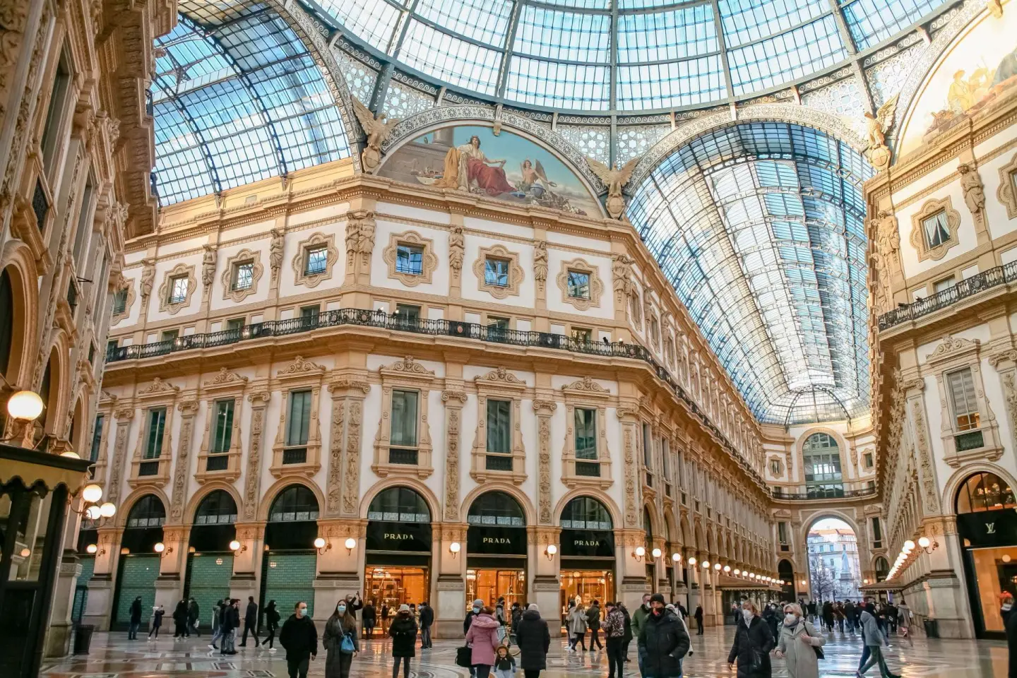 Interior de una galería comercial con una gran bóveda de cristal y hierro, fachadas de color crema con adornos dorados, pinturas en los arcos y tiendas iluminadas, con gente paseando por el suelo de mosaico