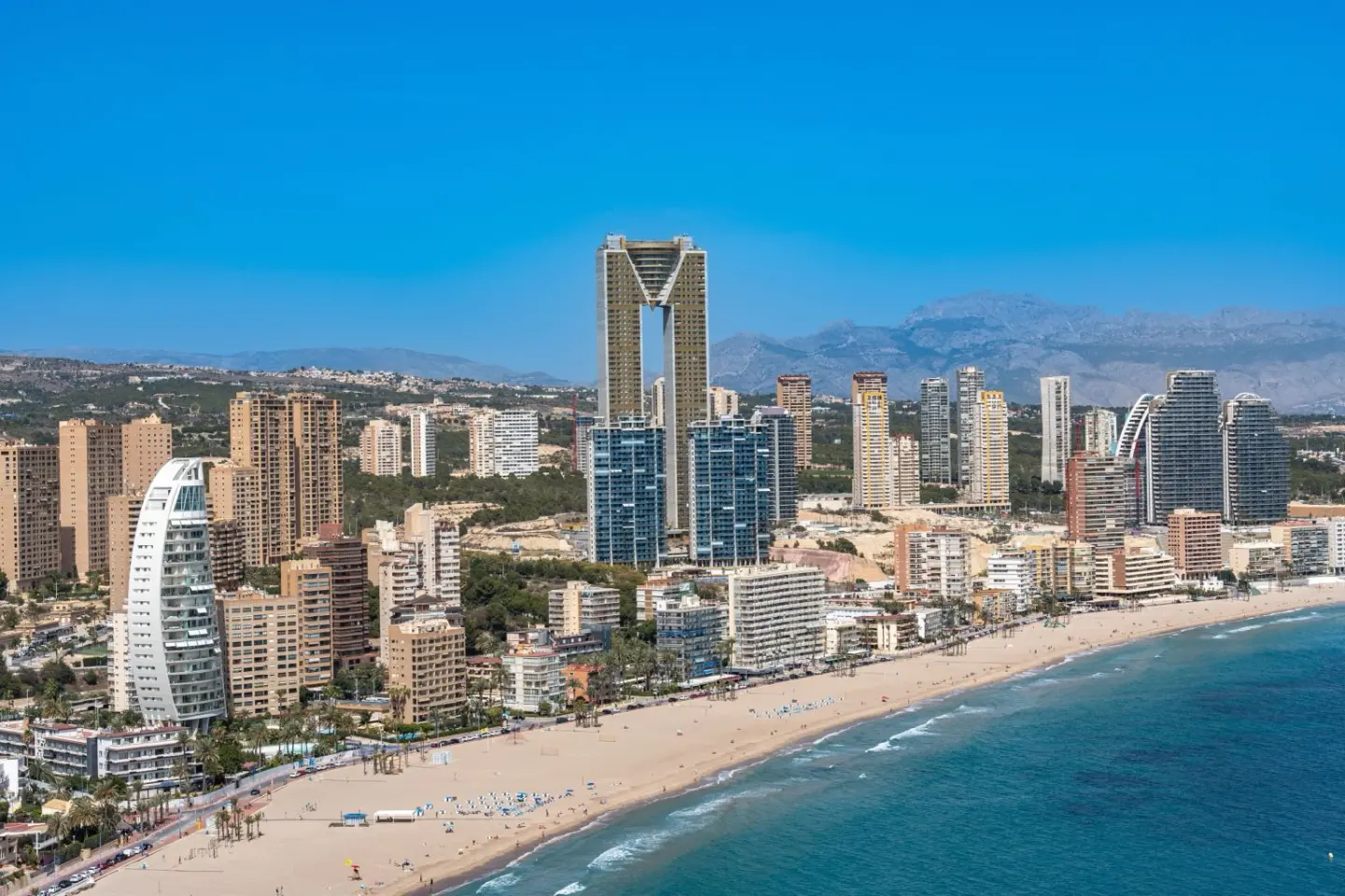 Rascacielos de una ciudad costera junto a una larga playa de arena, con montañas al fondo bajo un cielo azul