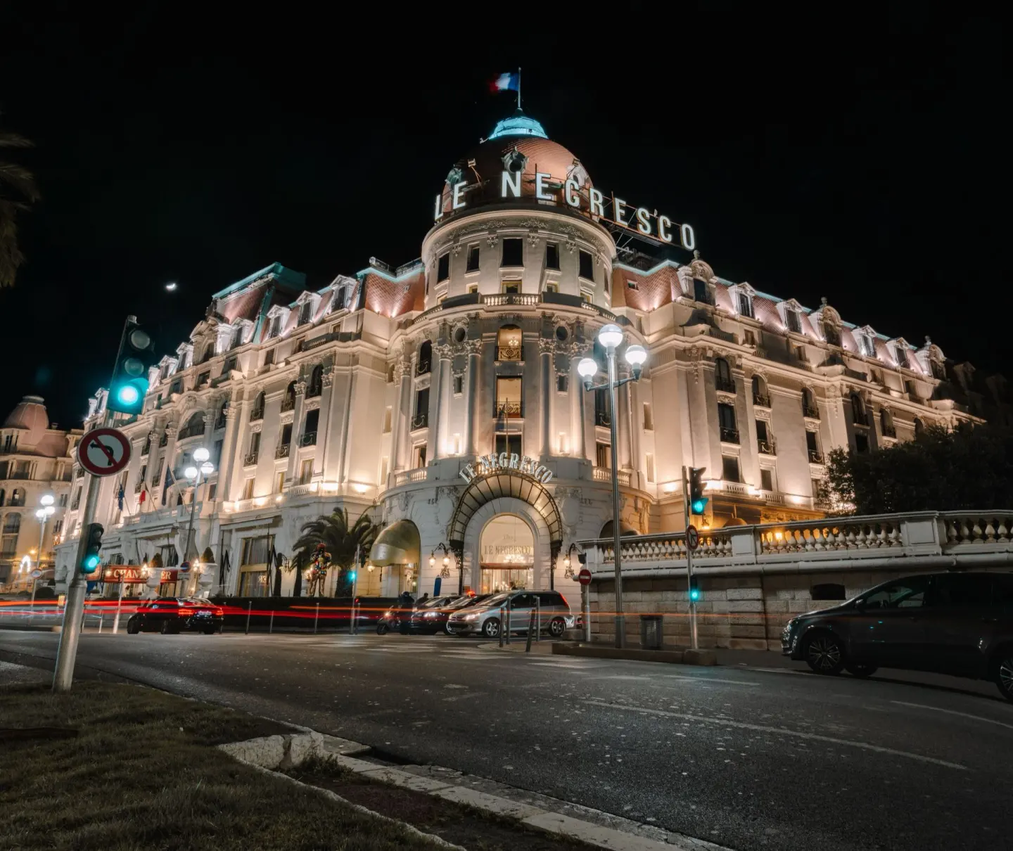 Gran hotel de estilo clásico con cúpula iluminado de noche, con el nombre «Le Negresco» en letras luminosas en lo alto, una bandera de Francia y estelas de luz de coches en la calle