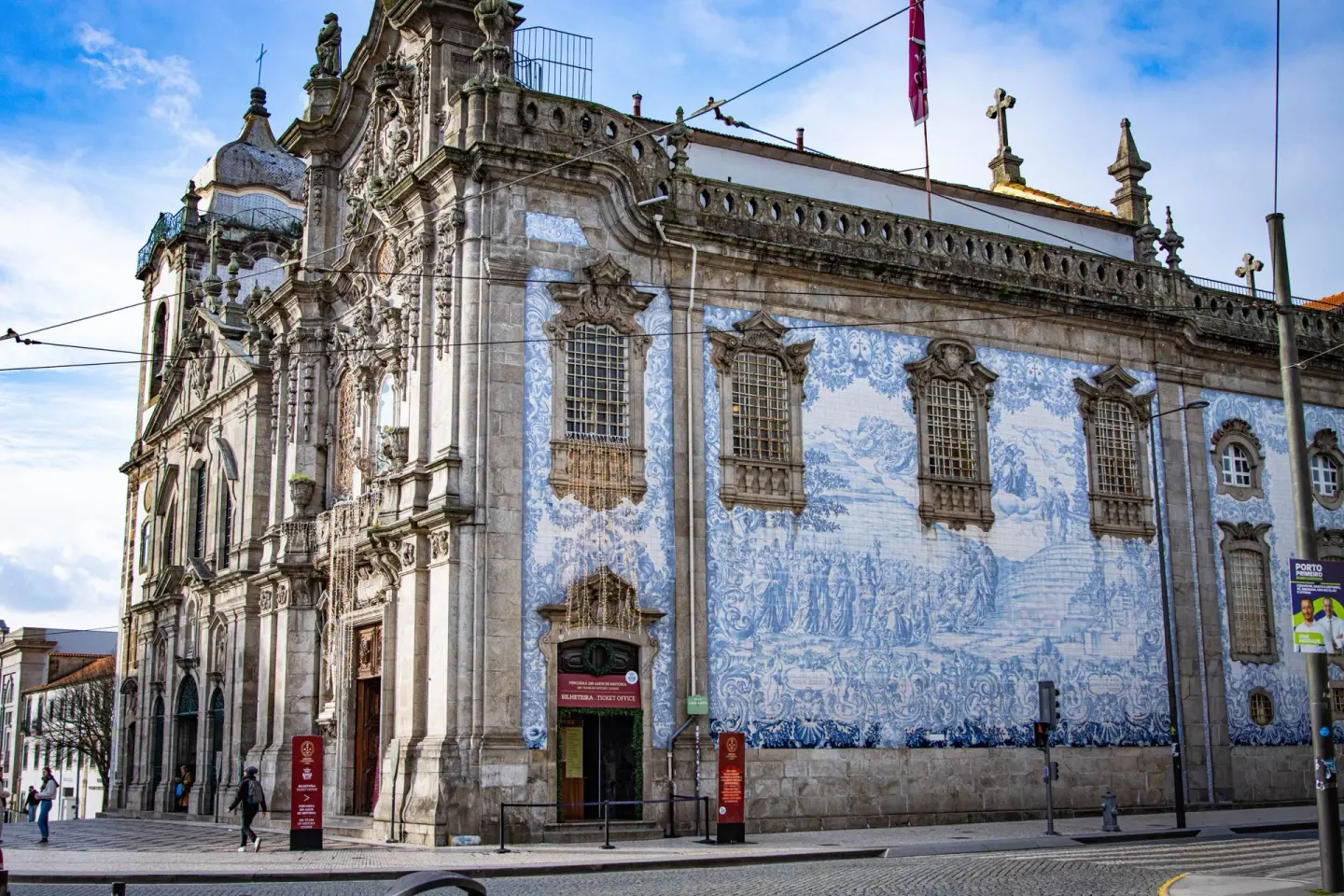 Iglesia barroca de granito con una gran fachada tallada y un lateral completamente cubierto de azulejos azules y blancos con una escena pintada y ventanas con marcos de piedra, en una esquina con gente caminando