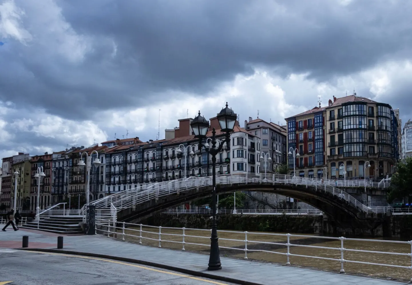 Puente peatonal blanco en arco sobre una ría, con edificios de fachadas con galerías acristaladas y un cielo cargado de nubes oscuras