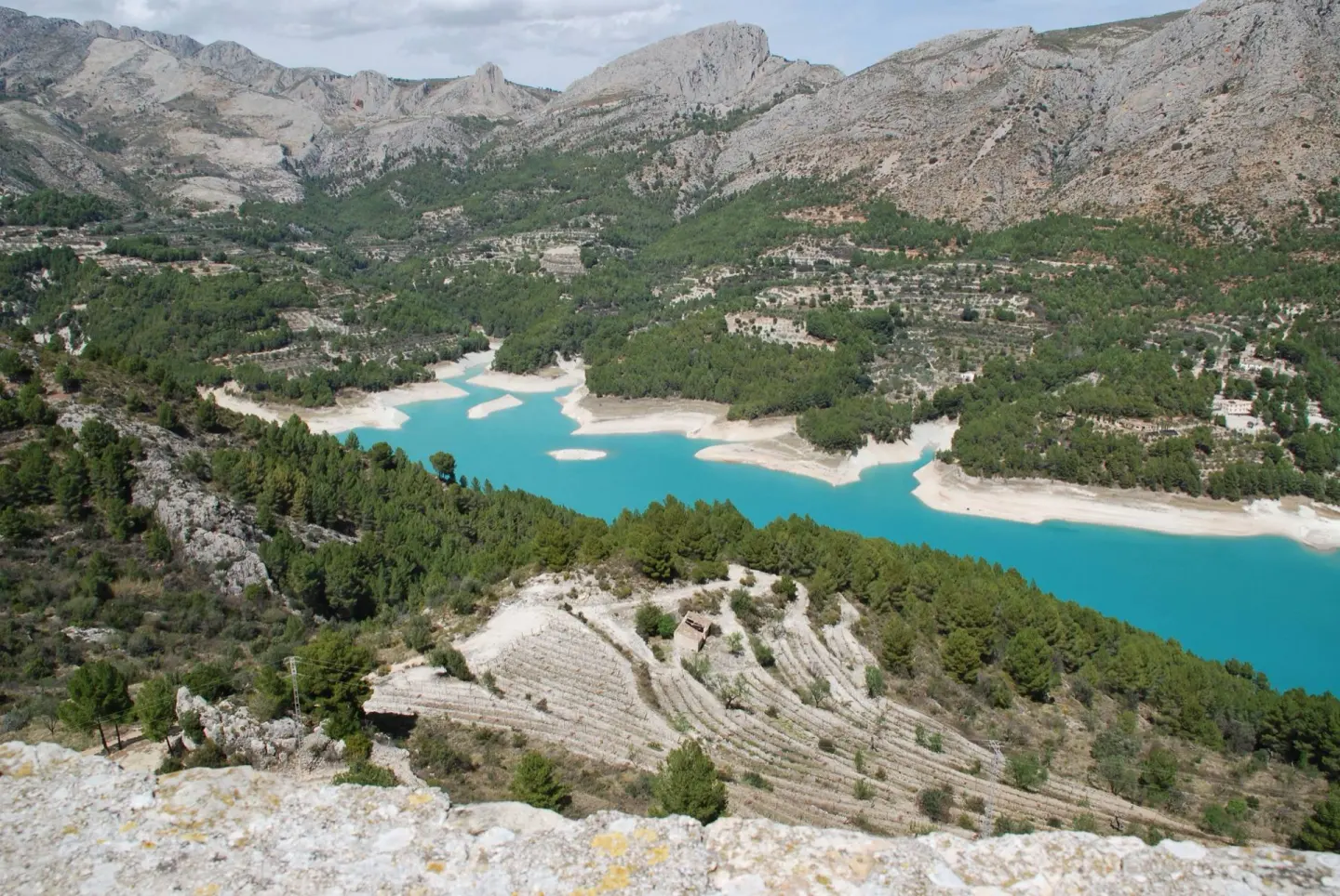 Embalse de aguas azul turquesa entre laderas de pinos y bancales, con montañas de roca al fondo y un muro de piedra en primer plano