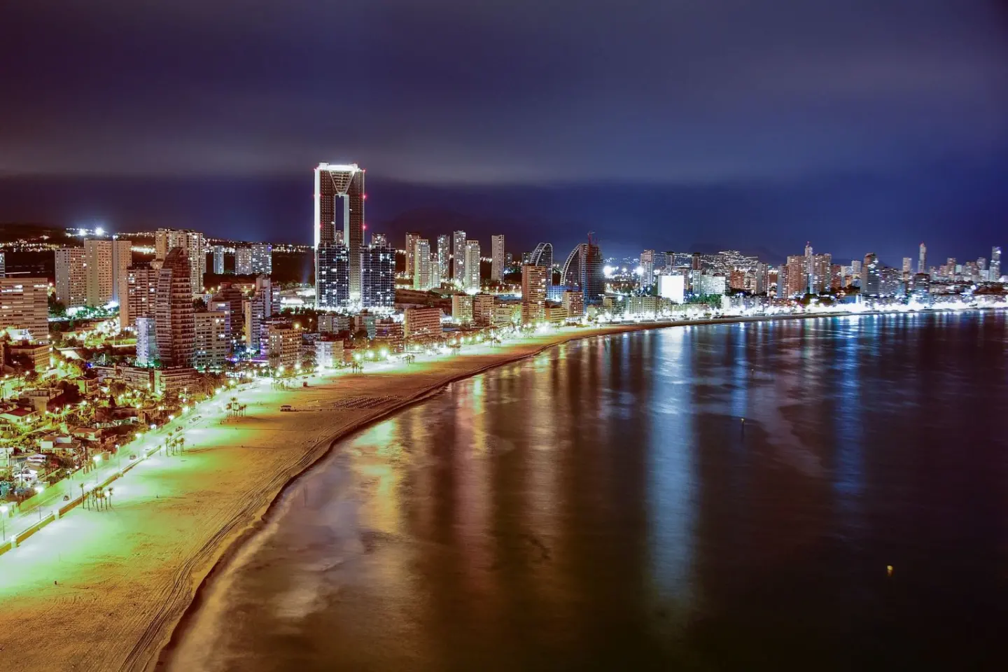 Vista nocturna de una playa y una ciudad de rascacielos iluminados, con el reflejo de las luces en el agua