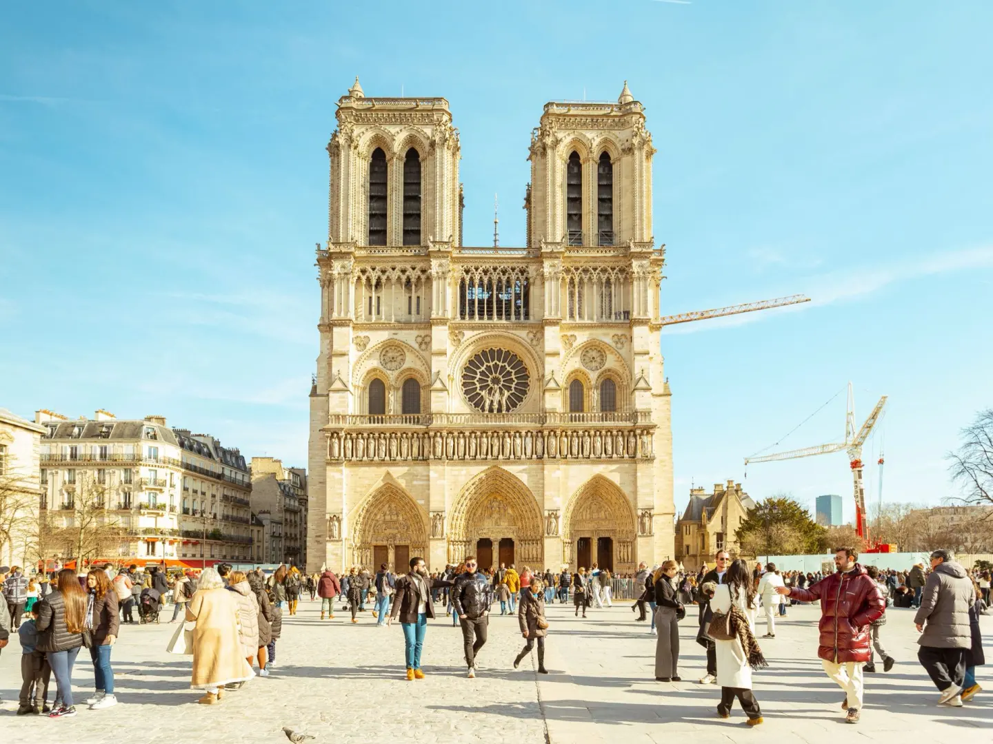 Fachada gótica de una catedral con dos torres cuadradas, un gran rosetón y tres portales, con una grúa de obra detrás y una gran explanada con mucha gente