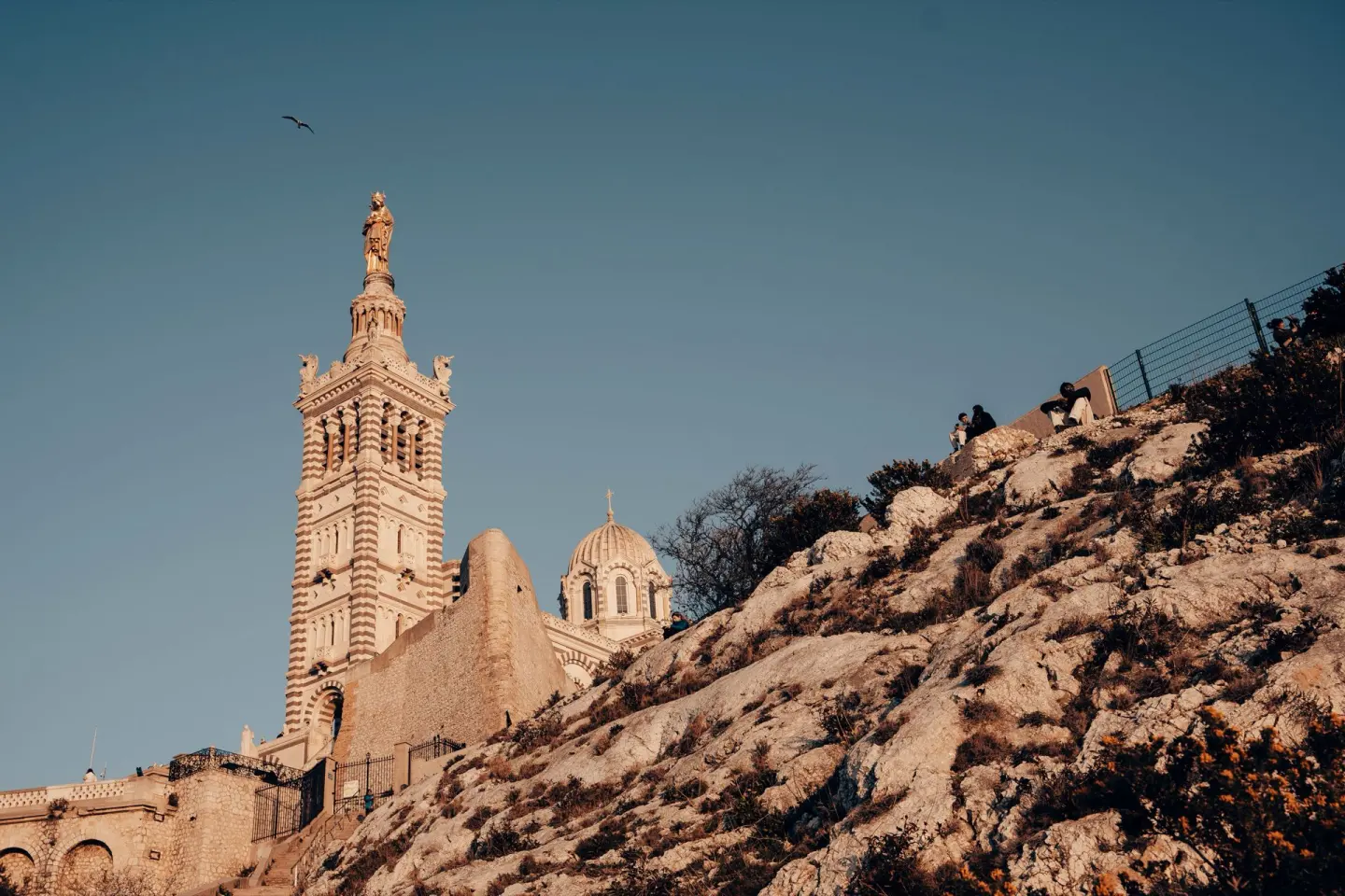 Basílica de torre alta con franjas de piedra clara y oscura y una estatua dorada en lo alto, sobre una ladera rocosa, con una gaviota en el cielo azul