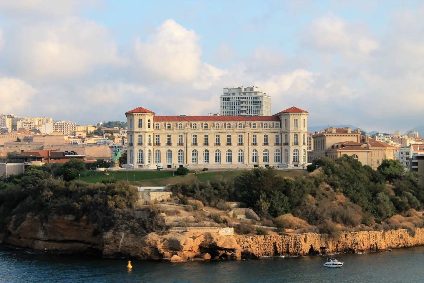 Gran palacio de piedra clara con tejados rojos y dos torres, sobre una colina verde junto al mar, con una lancha en el agua y la ciudad detrás