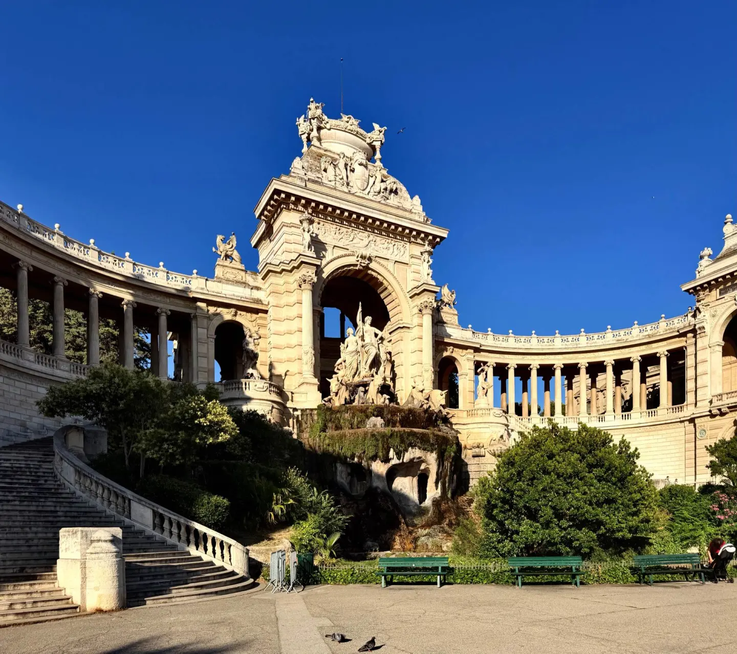 Monumento de piedra clara con una gran fuente de esculturas bajo un arco central y columnatas curvas a los lados, con una escalera, árboles y bancos verdes, bajo un cielo azul intenso