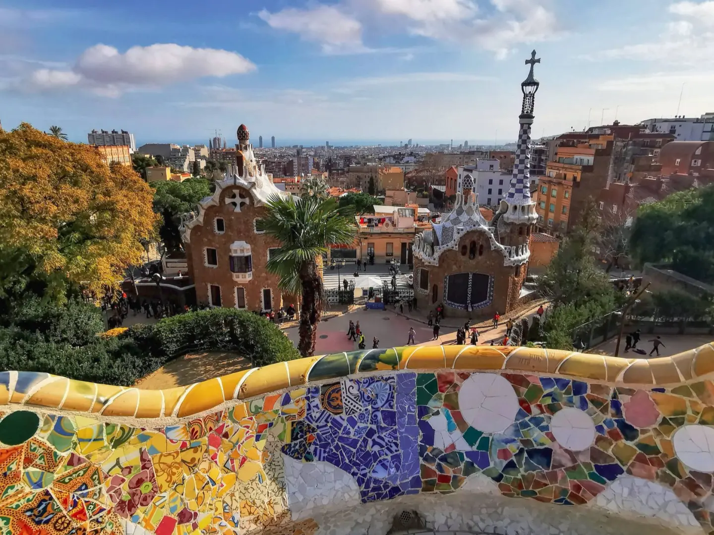 Banco ondulado de mosaico de colores en primer plano y, al fondo, dos casetas de fachada decorada y la ciudad hasta el mar
