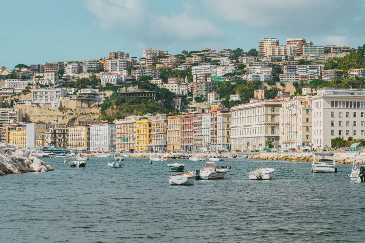 Fachada de edificios de colores amarillo, rosa y crema a lo largo de un paseo junto al mar, con una colina verde llena de casas detrás y barcas ancladas en el agua en primer plano