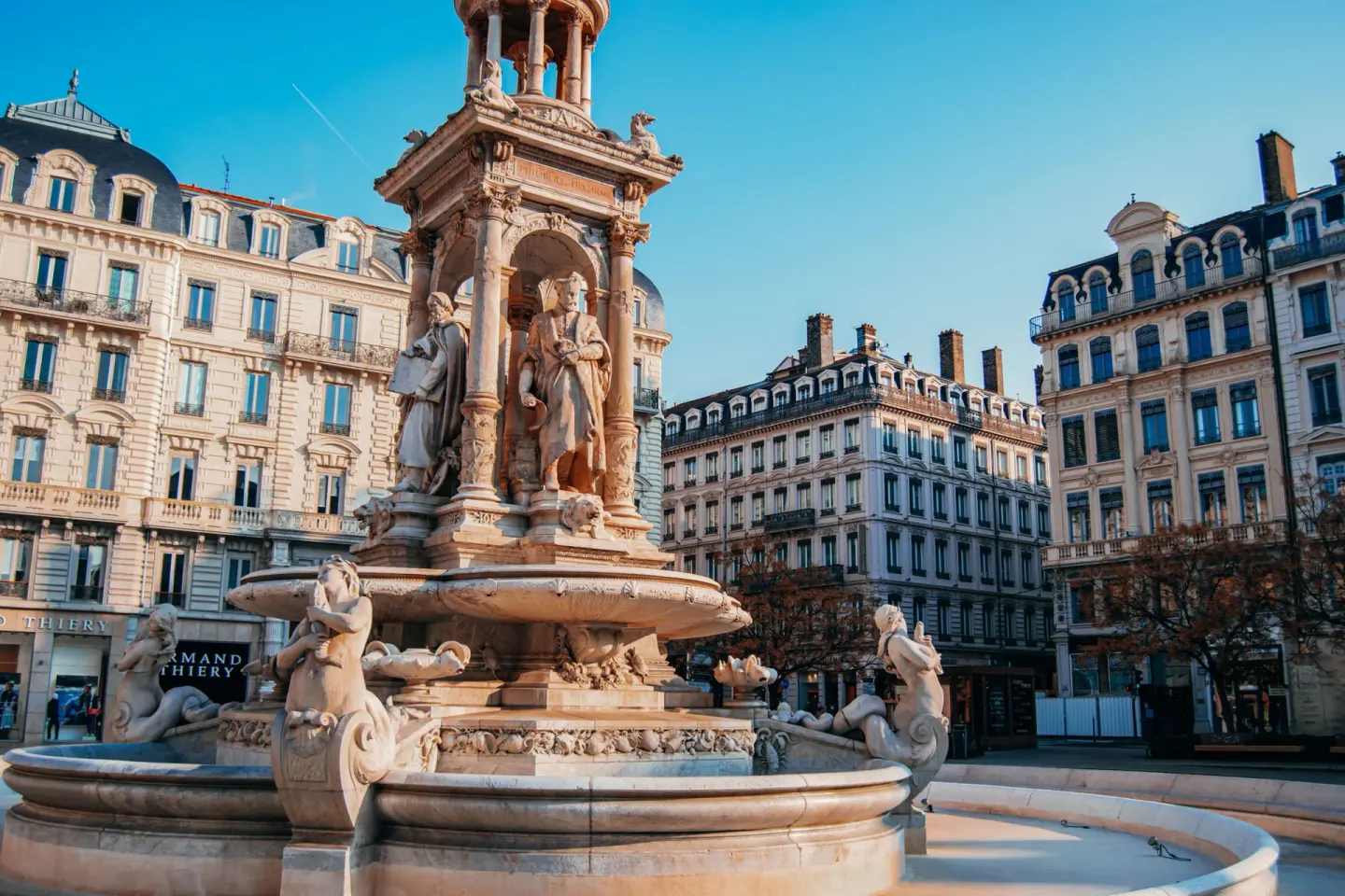 Fuente monumental de piedra con estatuas, figuras acuáticas y un templete en lo alto, en una plaza rodeada de edificios clásicos con tejados de pizarra, bajo un cielo azul
