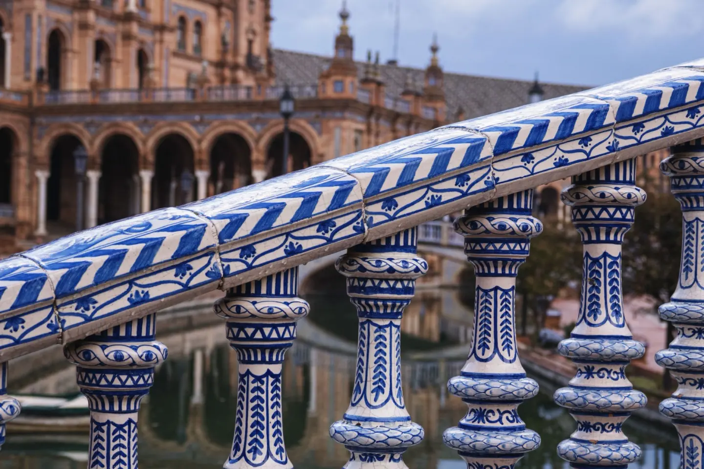 Barandilla de cerámica con dibujos azules sobre fondo blanco y balaustres decorados, con un canal y un edificio de ladrillo con arcos desenfocados al fondo