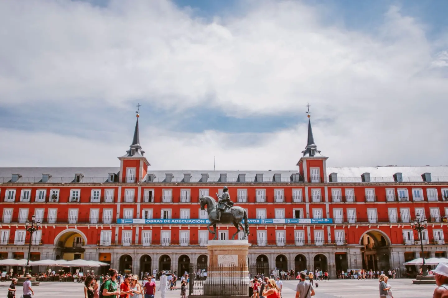 Gran plaza con edificios de fachada roja y balcones blancos, dos torres con chapiteles de pizarra, una estatua ecuestre de bronce y gente paseando