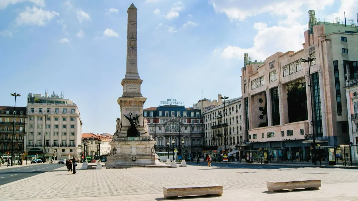 Plaza amplia con un alto obelisco de piedra con esculturas en la base, rodeada de edificios clásicos y de un gran edificio rosado de estilo años treinta, bajo un cielo azul con nubes