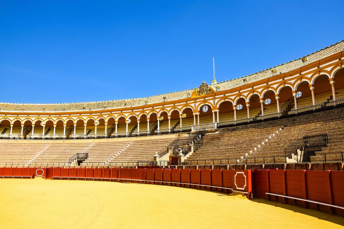 Interior de una plaza de toros con una galería de arcos amarillos y blancos sobre las gradas, una barrera roja y la arena amarilla, bajo un cielo azul intenso