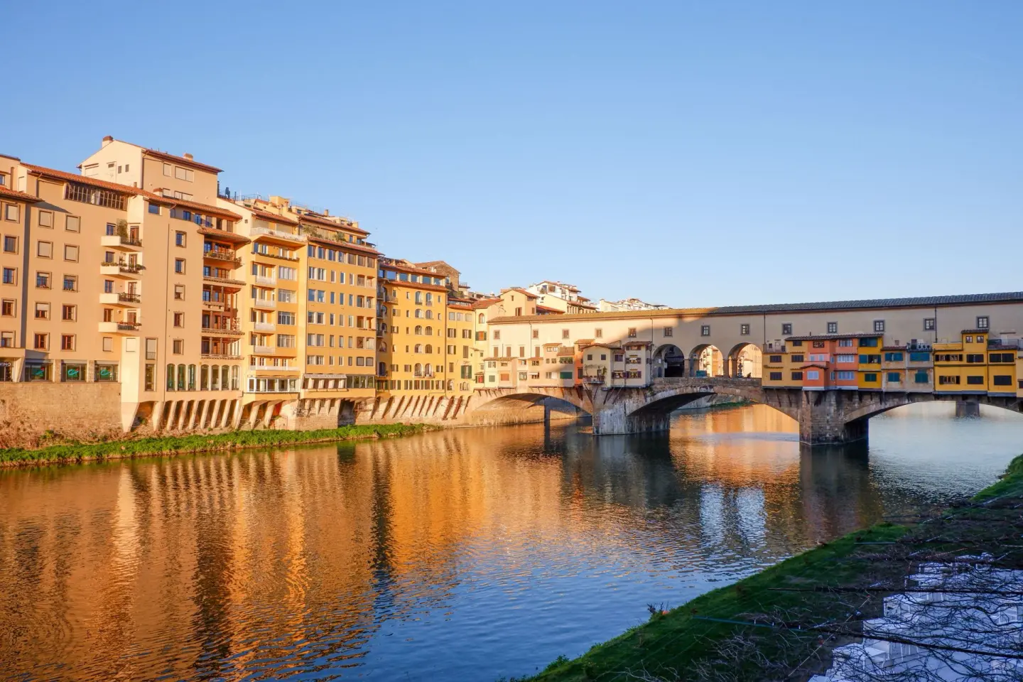 Puente de piedra de tres arcos sobre un río de aguas en calma, con casas de colores amarillo y naranja en su calzada, y edificios de fachadas ocres reflejados en el agua al atardecer