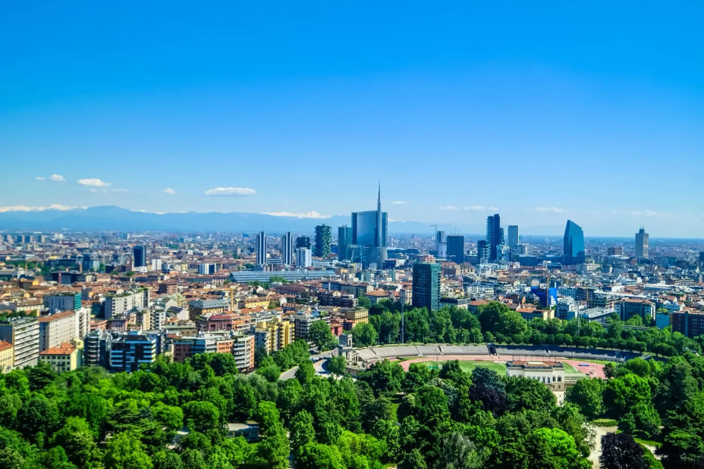 Vista panorámica de una ciudad desde lo alto, con rascacielos modernos de cristal al fondo, un gran parque de árboles verdes con una pista deportiva en primer plano y montañas lejanas bajo un cielo azul
