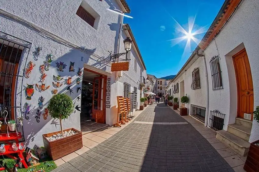 Calle peatonal de casas blancas con una tienda de artesanía que cuelga figuras de cerámica en la fachada, macetas y gente al fondo bajo un sol brillante