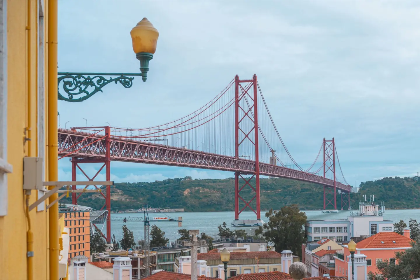 Gran puente colgante rojo sobre un río, visto entre un farol de forja verde y una fachada amarilla en primer plano, con tejados de teja y una colina verde al fondo