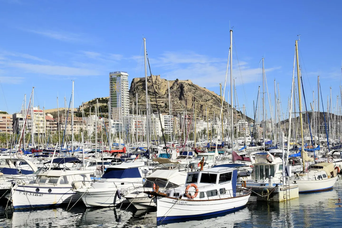 Puerto deportivo lleno de veleros y barcos de motor, con un monte rocoso coronado por una fortaleza y edificios de la ciudad al fondo