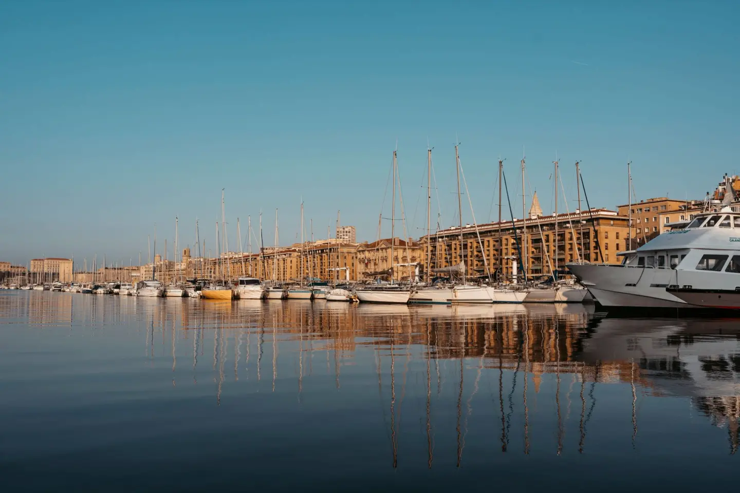Puerto lleno de veleros con sus mástiles reflejados en el agua en calma, y edificios de piedra dorada detrás bajo un cielo azul