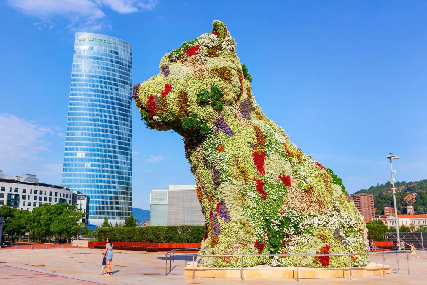 Gran escultura de un cachorro cubierta de flores de colores, con una torre de cristal azul al fondo y una mujer con un niño en primer plano