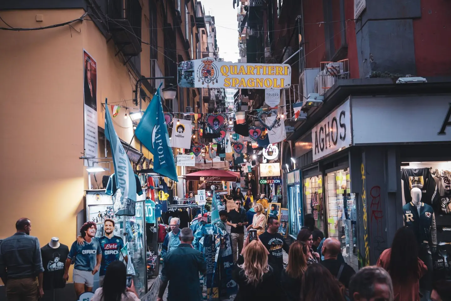 Calle estrecha y muy animada, llena de gente, con puestos y tiendas de camisetas y banderas de fútbol, un cartel sobre la calle con el nombre del barrio y fachadas de colores cálidos