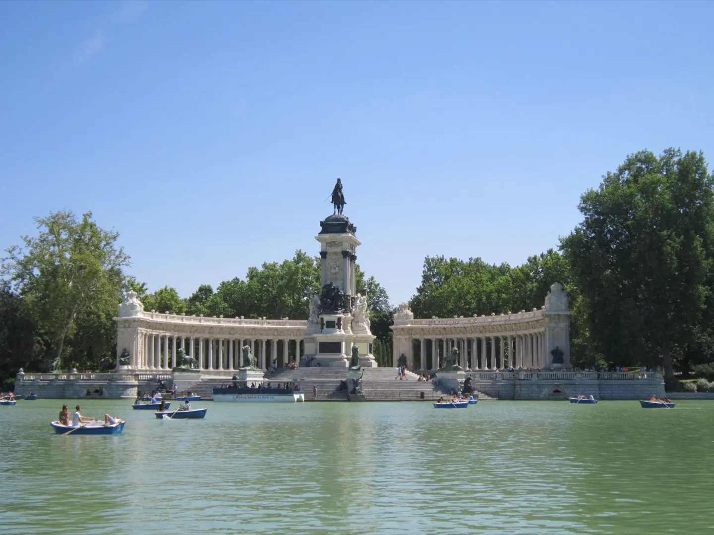Estanque con barcas de remos frente a un gran monumento con una columnata curva de piedra blanca, una escultura ecuestre y árboles detrás
