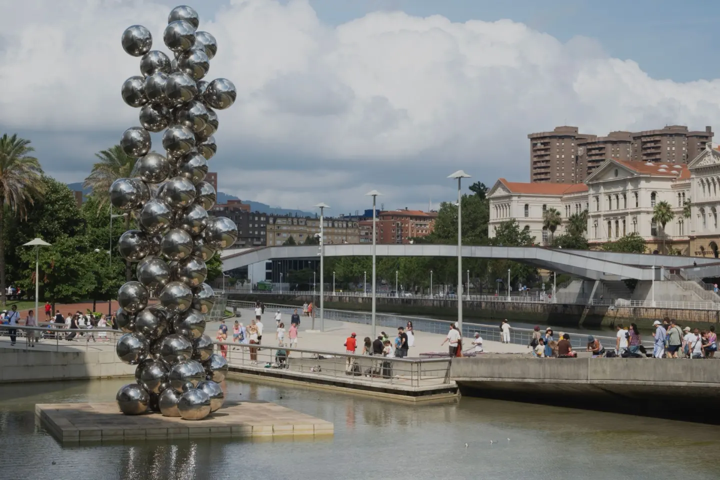 Escultura de grandes esferas plateadas sobre una plataforma en un estanque, con gente paseando junto a la ría y un edificio de tejados rojos al fondo