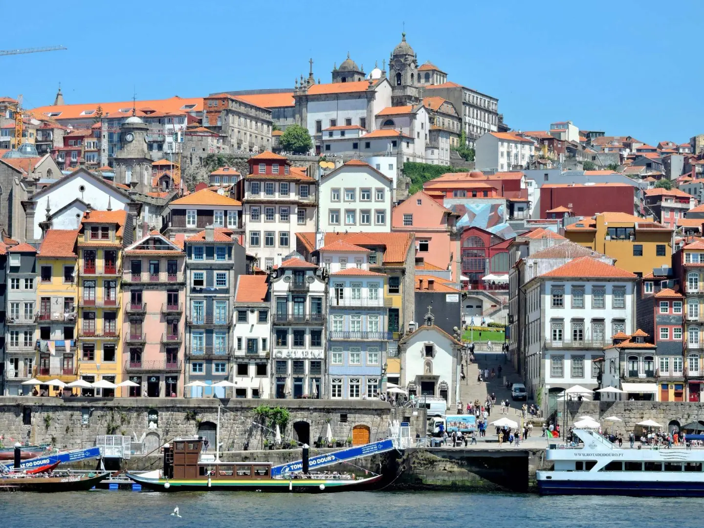 Fachadas de colores amarillo, rosa, blanco y azul con balcones y tejados de teja junto a un muelle de piedra, con terrazas con sombrillas blancas, barcos tradicionales de madera y un barco turístico en el agua, y una iglesia en lo alto de la colina