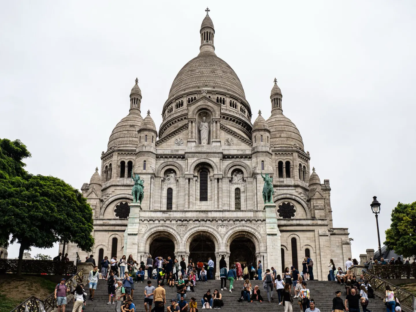 Basílica blanca con una gran cúpula y varias cúpulas menores, con dos estatuas ecuestres y una escalinata llena de gente en primer plano, bajo un cielo nublado