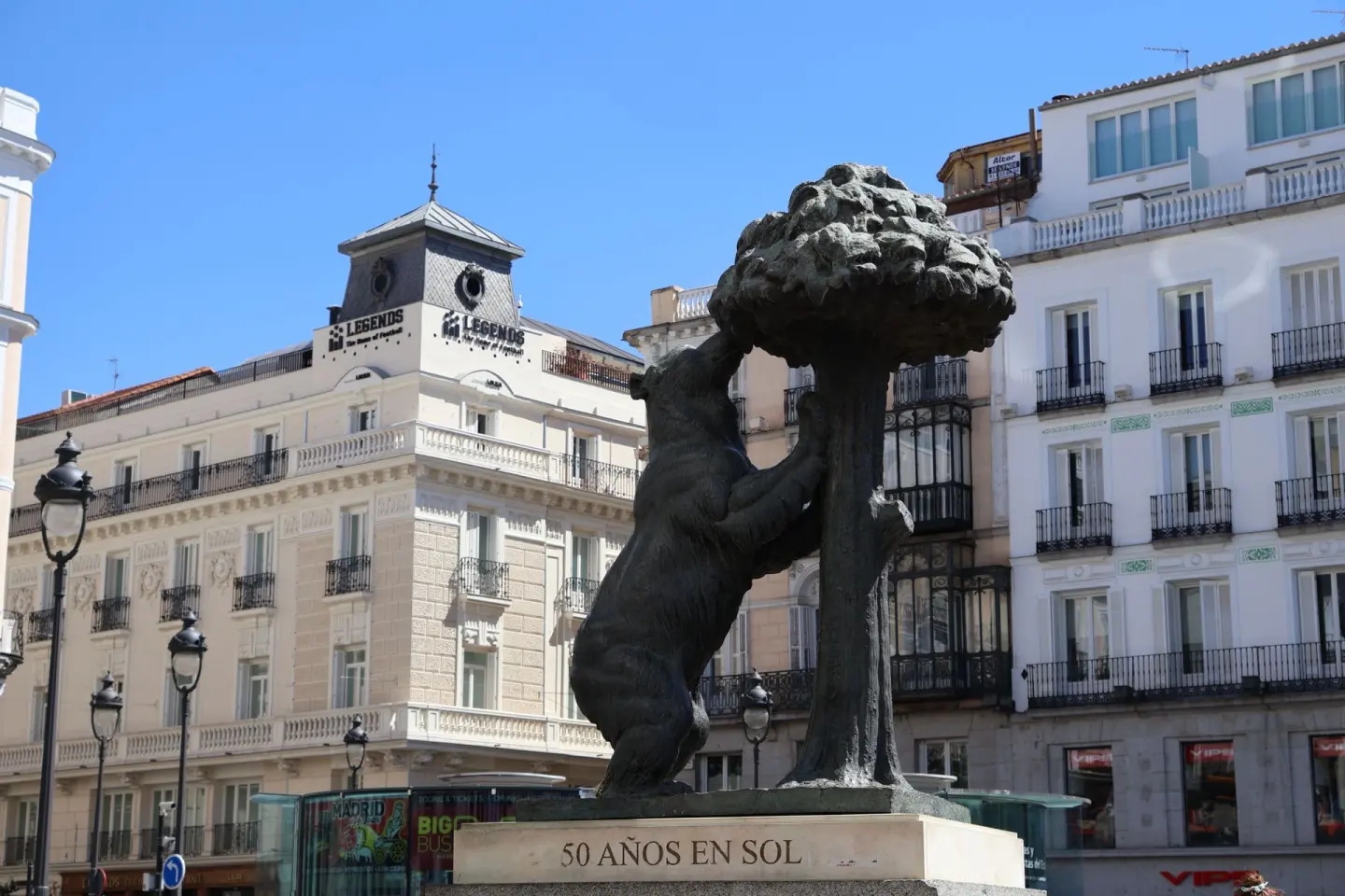 Escultura de bronce de un oso apoyado en un madroño, con edificios claros de balcones de hierro al fondo bajo un cielo azul
