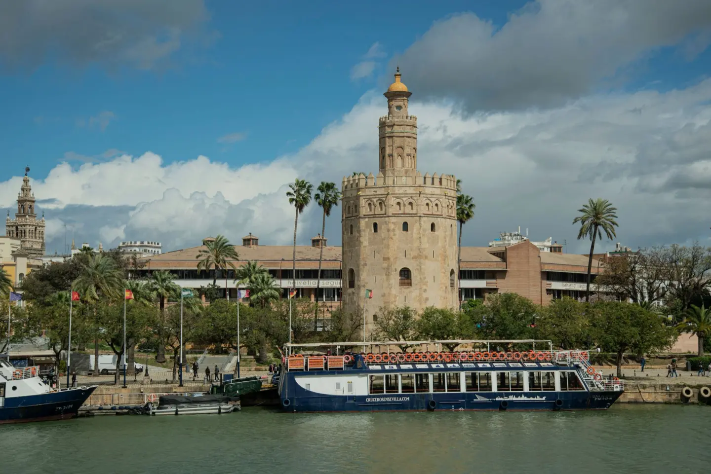 Torre de piedra de forma poligonal con remate dorado junto a un río, con palmeras y un barco turístico amarrado en primer plano